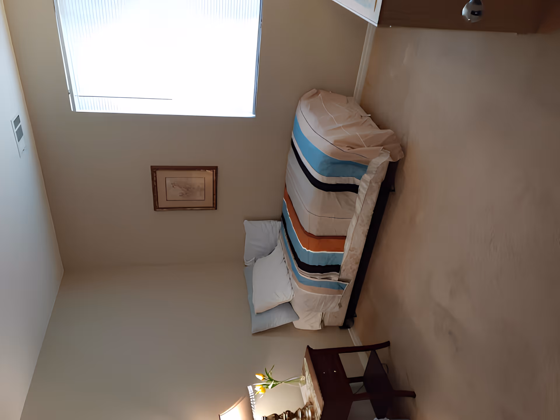 Small bedroom with a single bed dressed in striped bedding, a nightstand with a lamp and flowers, a framed picture on a beige wall and a window above.