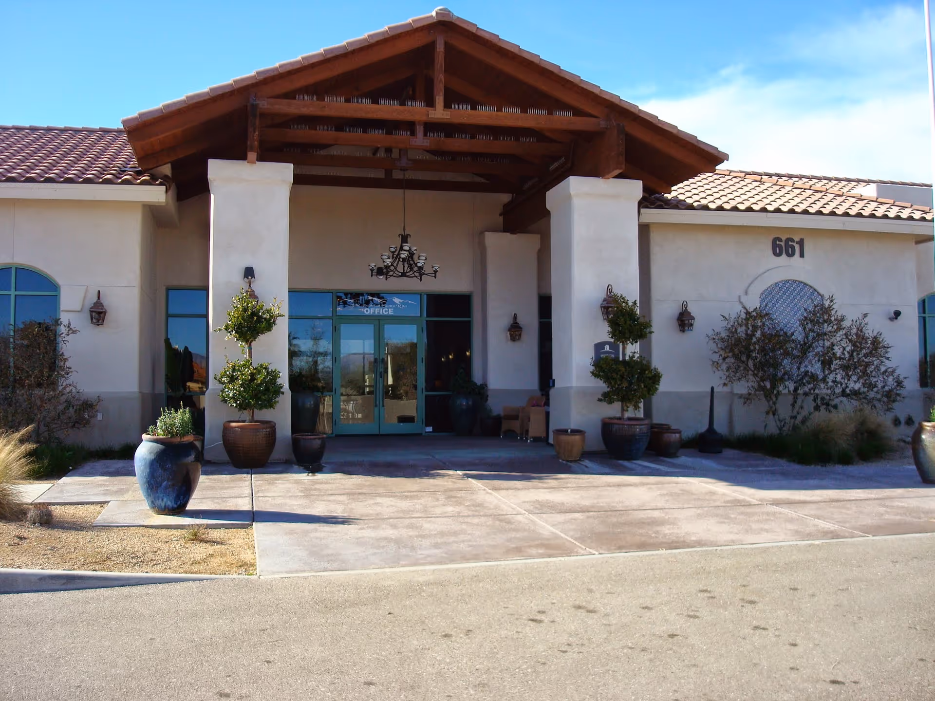 Front exterior view of a building with a covered entrance supported by two large columns. The building has a tiled roof and several potted plants near the entrance. The number 661 is displayed on the right side of the building above a decorative window. The entrance doors are glass with a sign that reads 'OFFICE'.