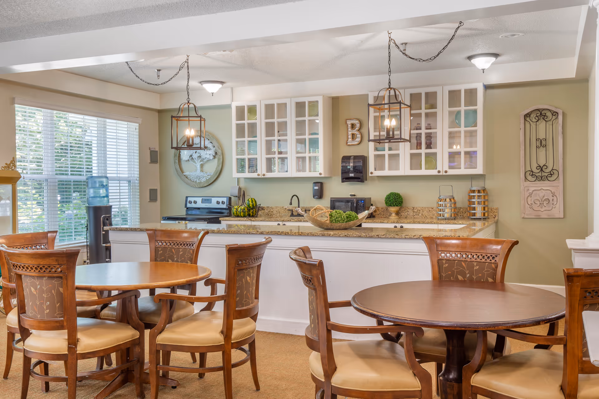 A bright and inviting dining area with round wooden tables and cushioned chairs. In the background, there is a kitchen with white cabinets, granite countertops, a stove, microwave, and decorative items. Large windows with blinds allow natural light to fill the space.