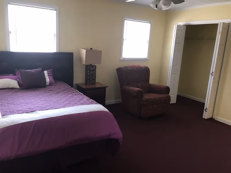 A bedroom with a purple bedspread, bedside table and lamp, an upholstered armchair, two windows, and an open closet.