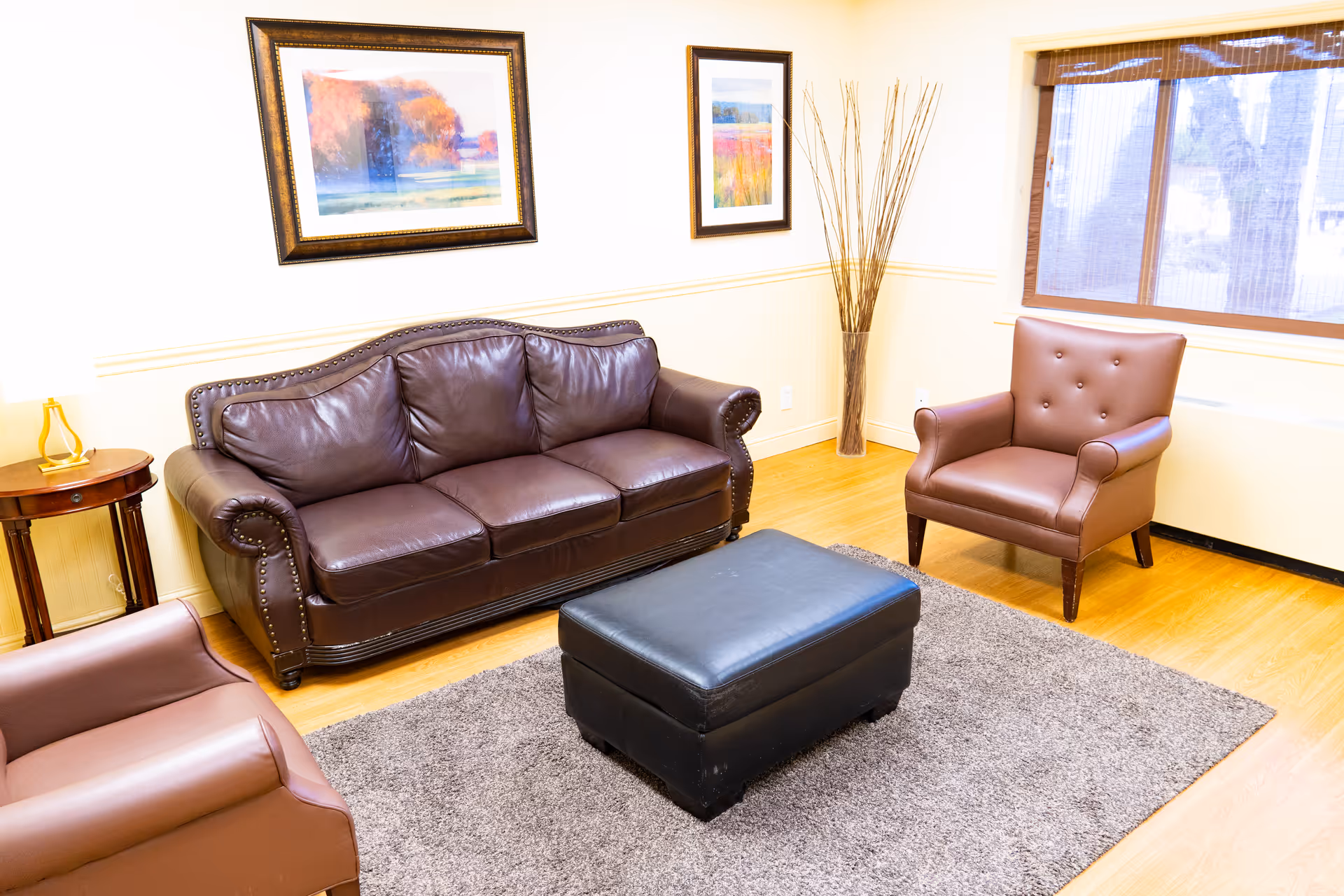 A cozy living room with a brown leather sofa, two matching brown leather armchairs, a black leather ottoman on a gray rug, a small wooden side table with a lamp, two framed paintings on the wall, and a window with wooden blinds letting in natural light.