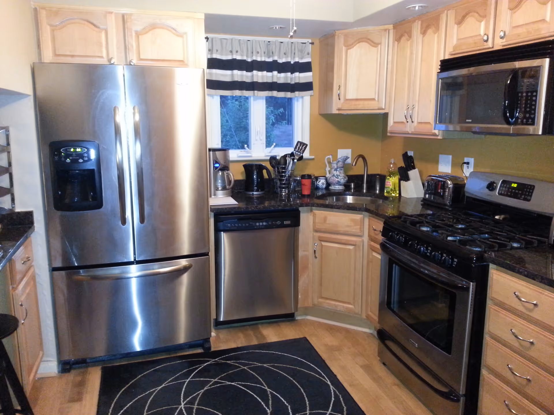 A modern kitchen with stainless steel appliances including a refrigerator, dishwasher, microwave, and gas stove. The kitchen has light wood cabinets, a black countertop, a small window with a black and white striped valance, and various kitchen utensils and items on the counter. A black rug with a white circular pattern is on the wooden floor.