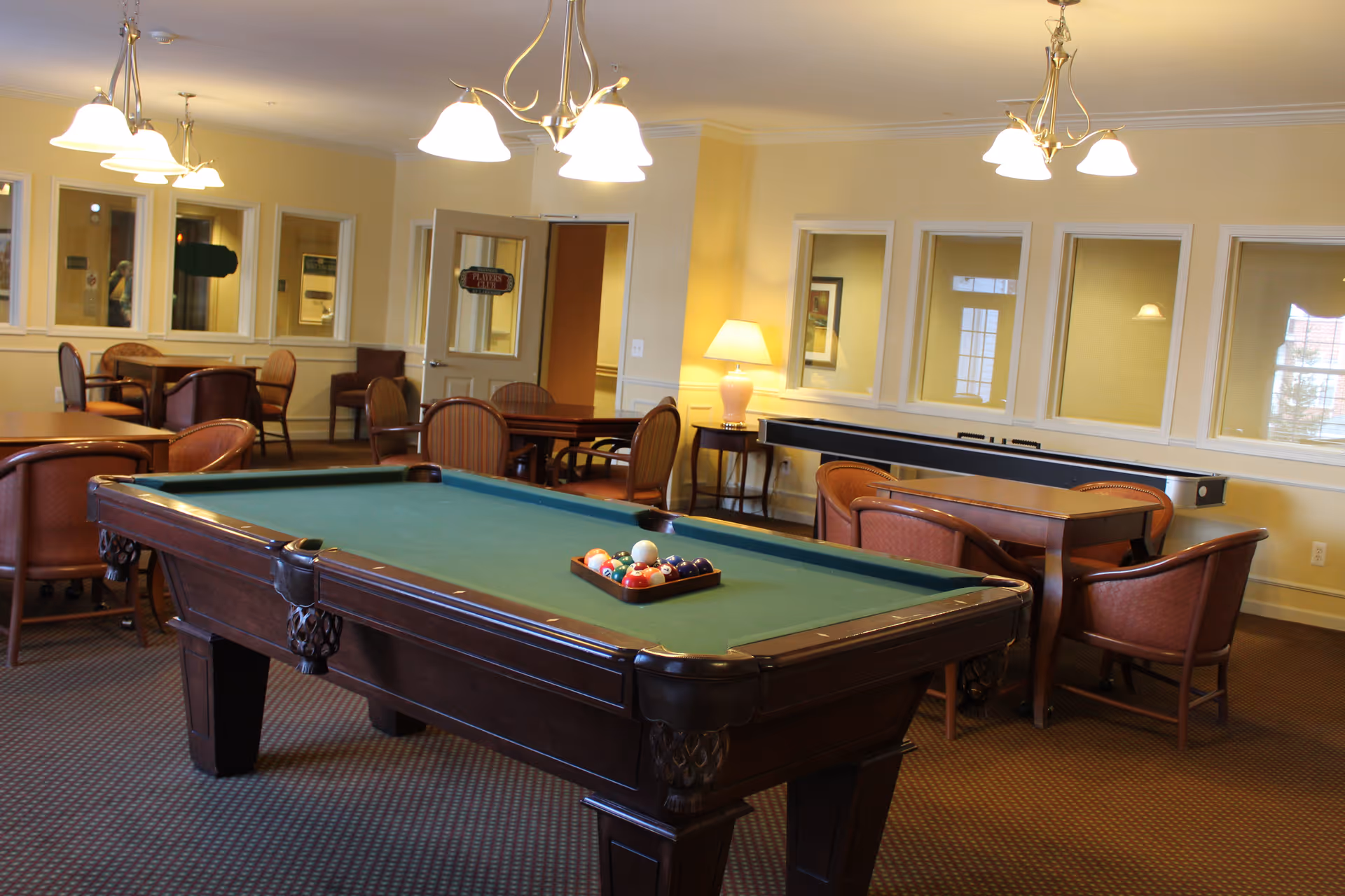 A well-lit recreational room featuring a pool table with balls arranged in a triangle, several tables with chairs around them, a shuffleboard table against the wall, and warm yellow walls with multiple windows and a lamp on a side table.