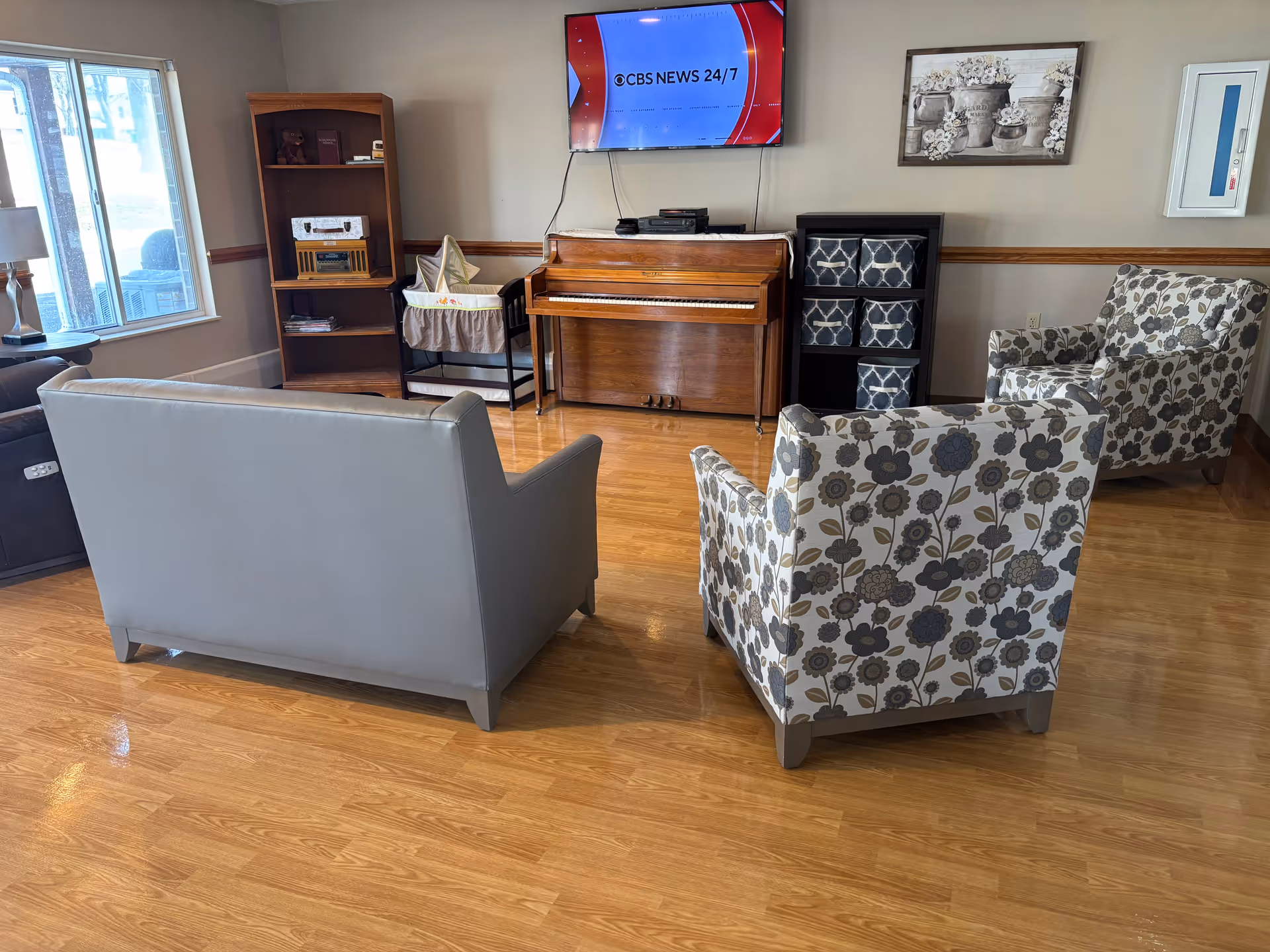 A cozy living room area with a wooden floor, featuring a gray sofa, two floral-patterned armchairs, a wooden piano against the wall, a TV mounted above the piano displaying CBS News 24/7, a wooden bookshelf with decorative items, a bassinet, and a small black storage unit with patterned bins. A window on the left side lets in natural light.