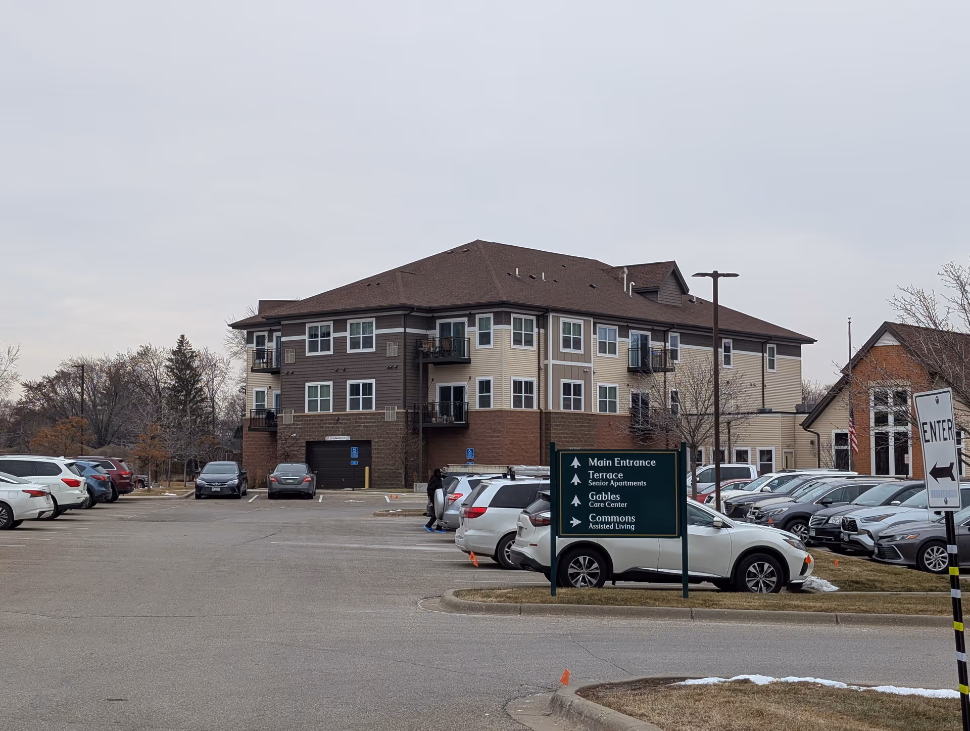 Parking lot with several cars in front of a multi-story senior living facility building with balconies. A green directional sign indicates the main entrance, terrace senior apartments, gables care center, and commons assisted living. Trees and a cloudy sky are visible in the background.