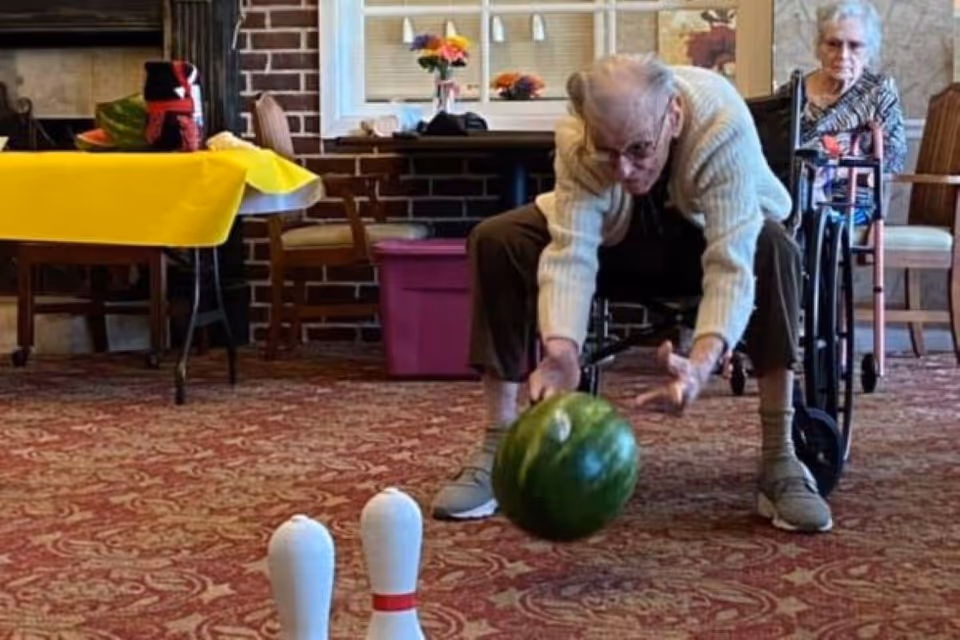 An elderly man in a wheelchair is rolling a green bowling ball towards two white bowling pins set up on a carpeted floor. Another elderly woman in a wheelchair watches in the background. The room has a brick wall, a window, and a table with a yellow tablecloth and some items on it.