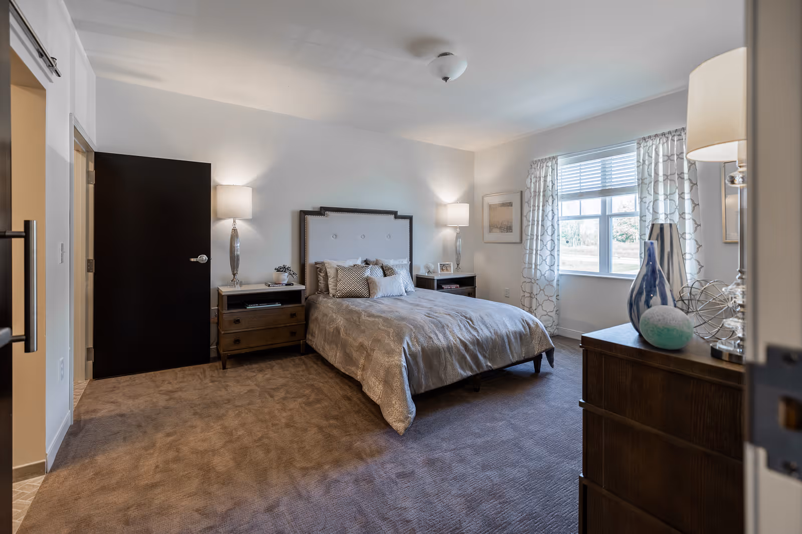 A well-lit bedroom with a large bed centered against the back wall, flanked by two nightstands each with a lamp. The bed has a tufted headboard and is covered with a beige comforter and multiple pillows. There is a window with patterned curtains on the right wall letting in natural light. A dresser with decorative items is visible in the foreground on the right side. The room has beige carpeting and neutral-colored walls.