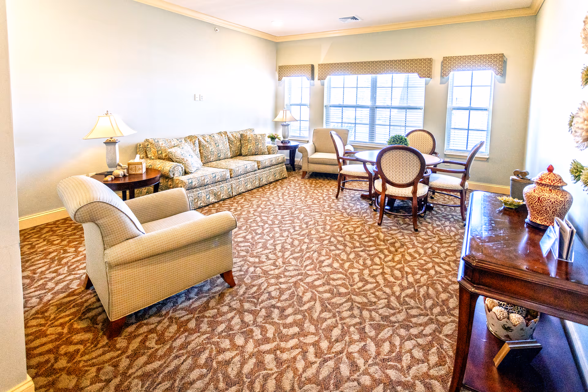 A cozy living room with patterned carpet, a floral-patterned sofa, an armchair, a round wooden table with four upholstered chairs, two table lamps on side tables, and a wooden console table with decorative items. Large windows with valances allow natural light to fill the room.