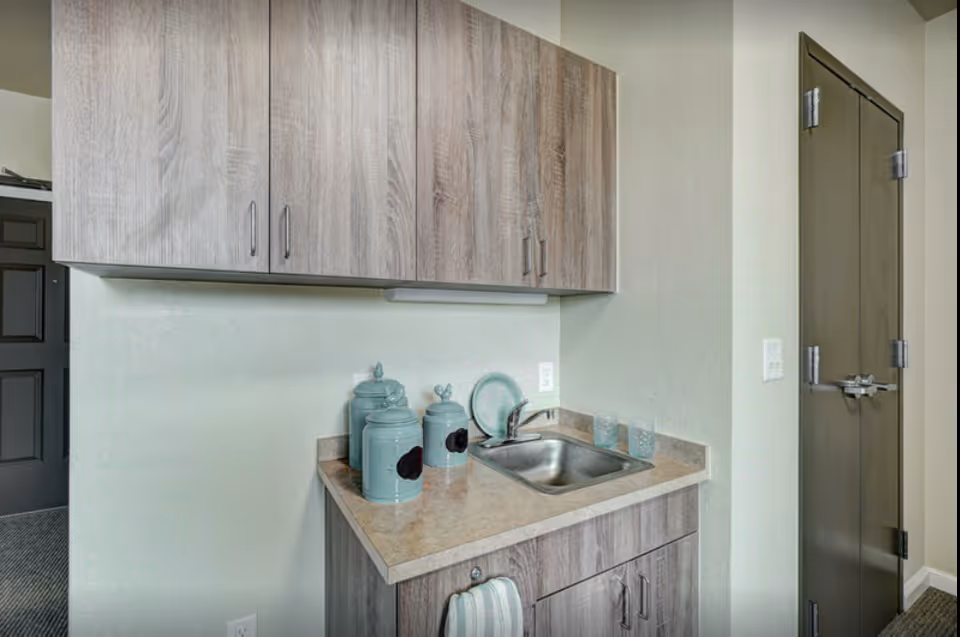 A small kitchenette area with a stainless steel sink, beige countertop, and light wood cabinets above and below. On the countertop are three teal canisters, a teal plate, and two clear glasses. A striped towel hangs from the cabinet handle below the sink. A closed dark-colored door is visible to the right.