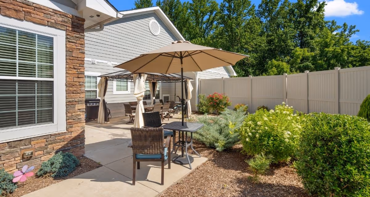 Sunny outdoor patio and garden area with umbrella-shaded table and chairs beside a fenced building.