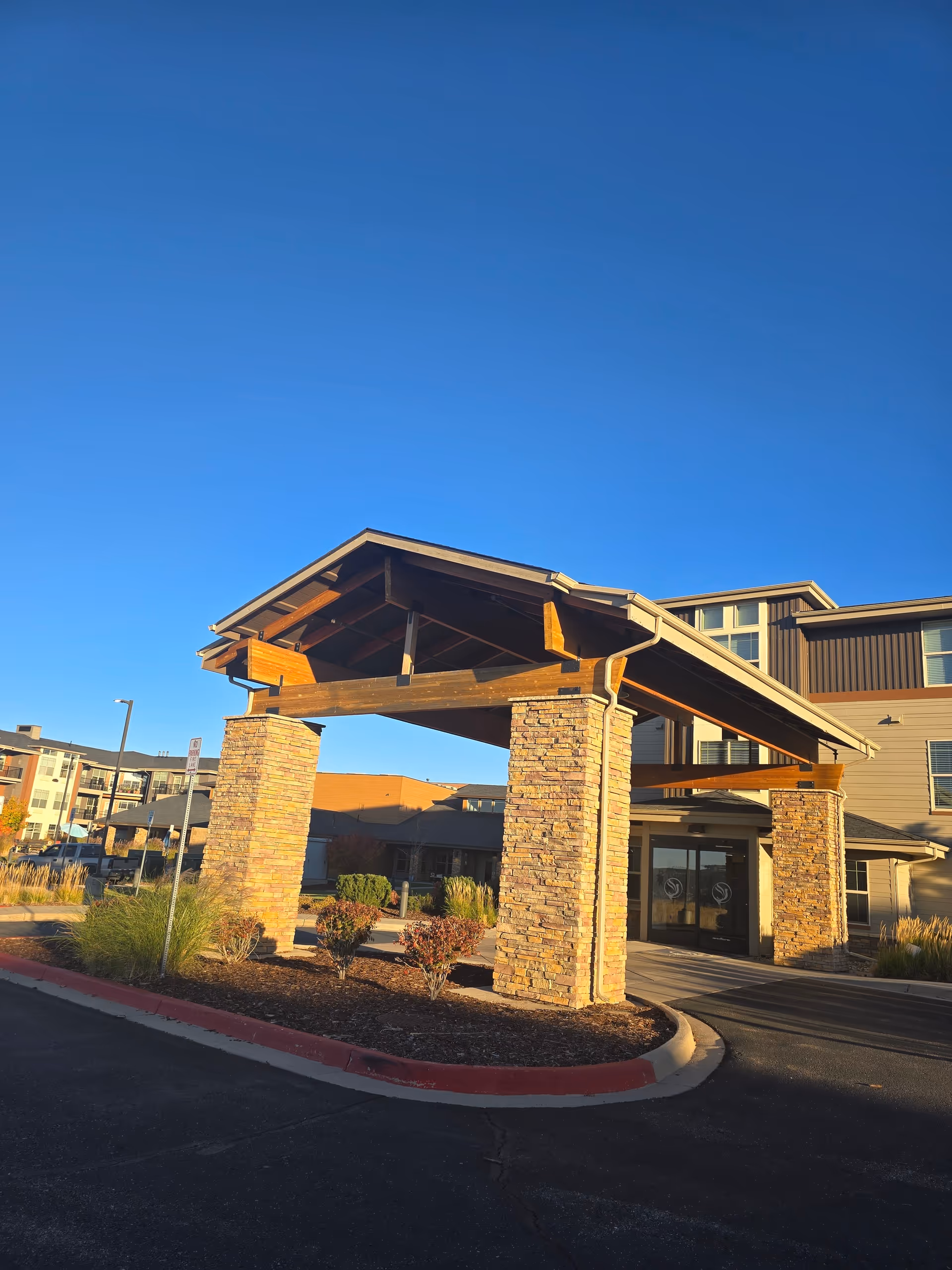 Entrance of a senior living facility with a covered driveway supported by stone pillars, surrounded by landscaping and a clear blue sky.