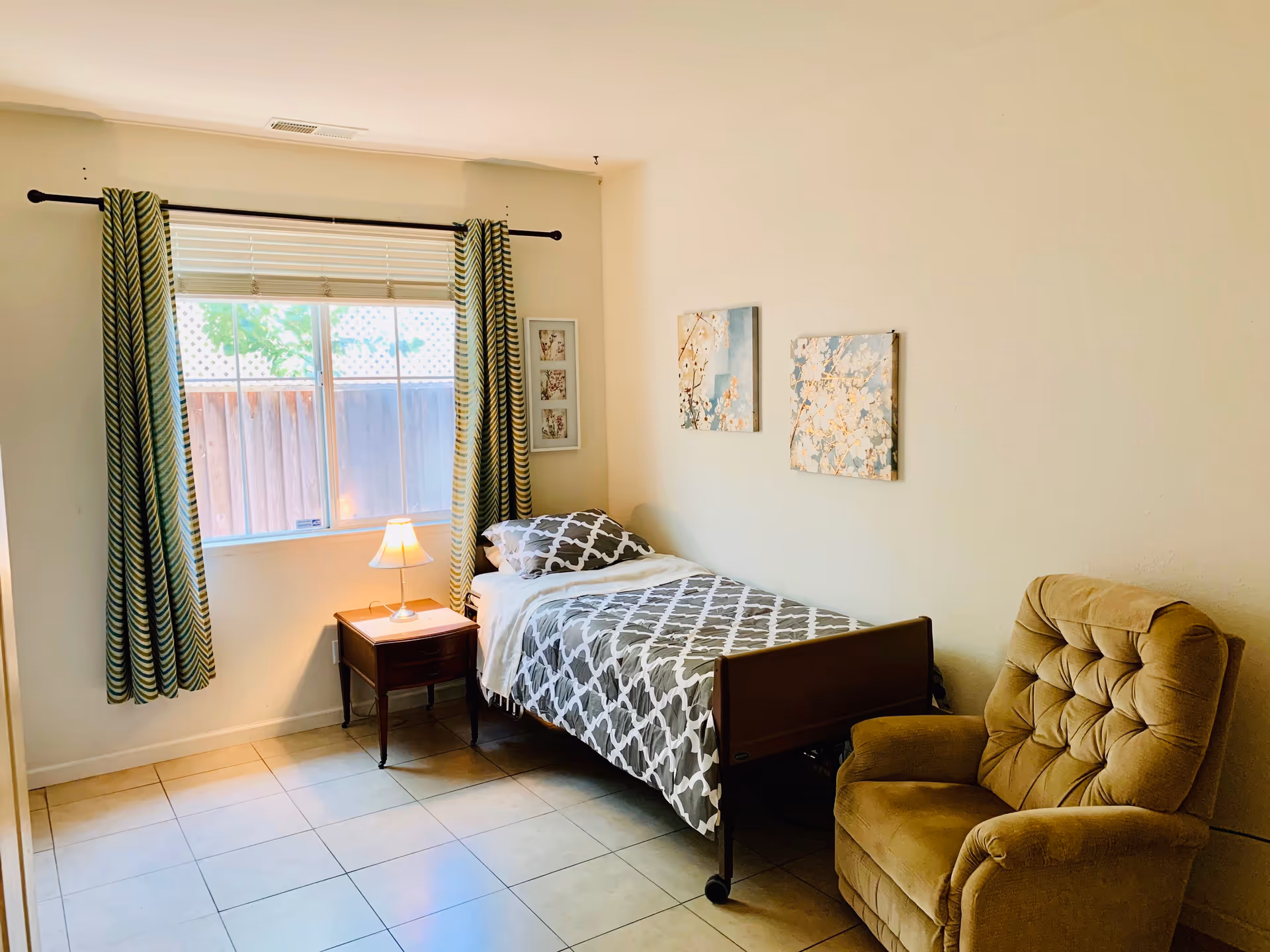 A simple elderly care bedroom with a single bed covered in a gray and white patterned blanket, a wooden nightstand with a small lamp, a large window with green and white striped curtains, two floral paintings on the wall, and a cushioned beige armchair.