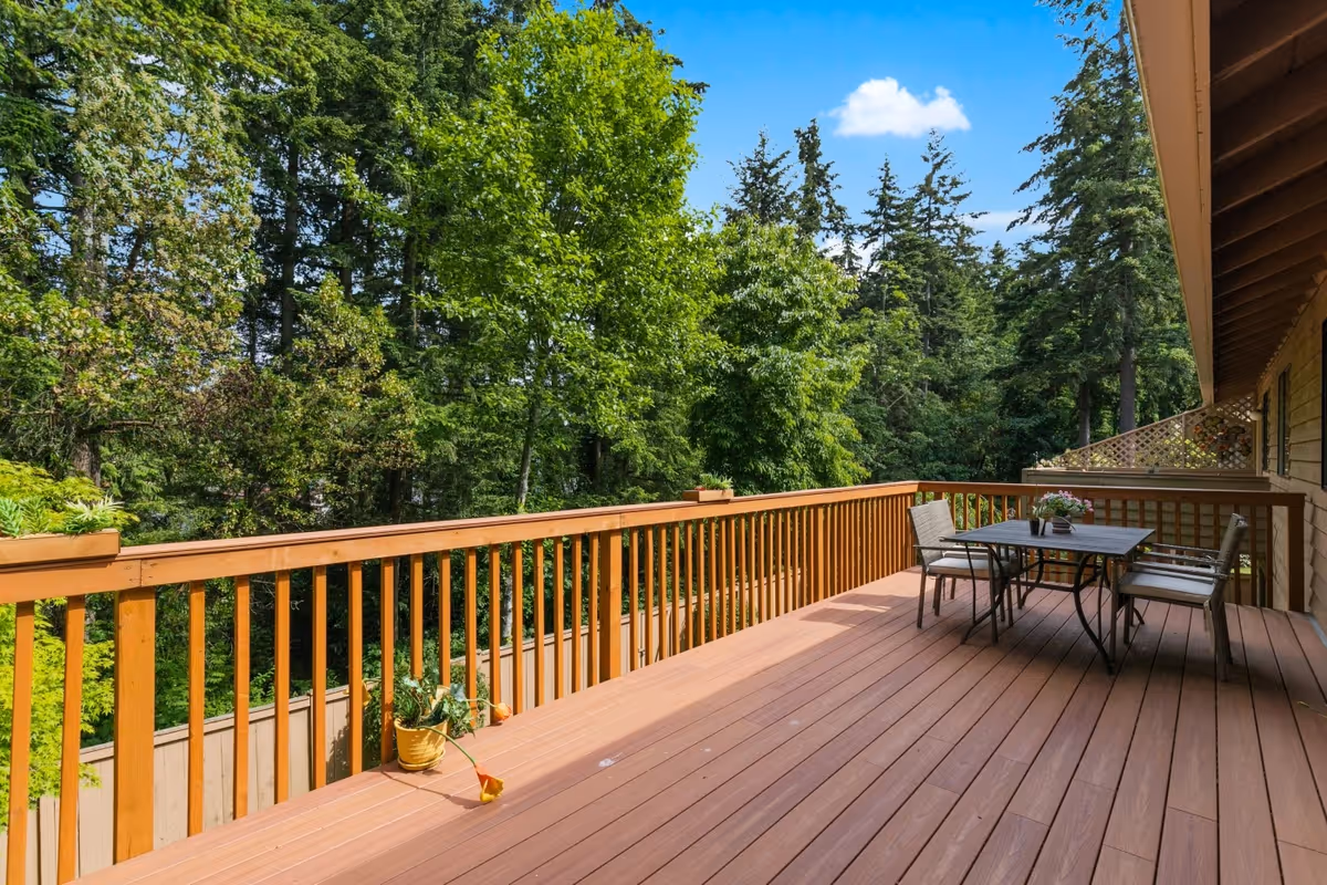 A spacious wooden outdoor deck with a table and four chairs, surrounded by lush green trees under a clear blue sky.