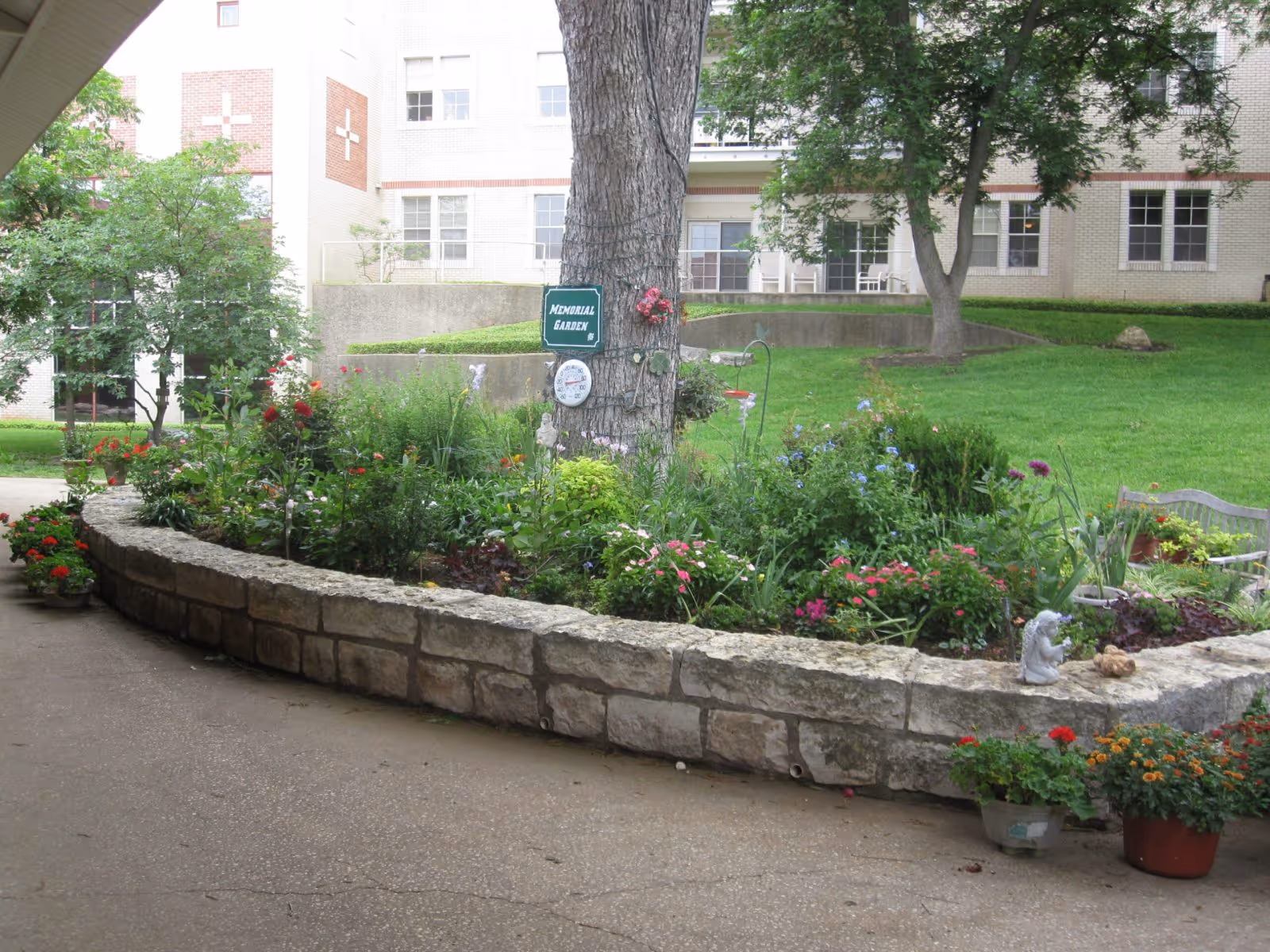 A garden area with a stone border filled with various flowering plants and greenery. A large tree stands in the middle with a sign that reads 'Memorial Garden' and a thermometer attached to it. In the background, there is a multi-story brick building with windows and some outdoor seating visible on a patio.