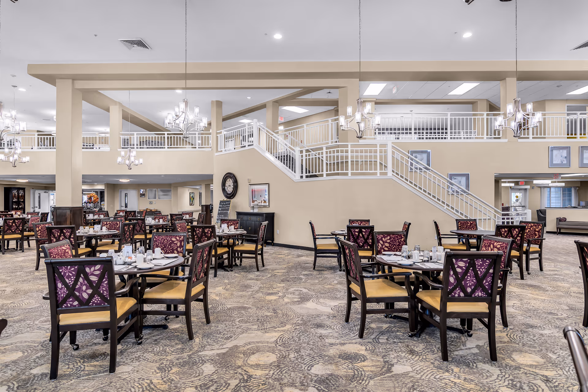 Spacious dining room with multiple round tables and chairs featuring purple patterned backs and yellow seats. The room has beige walls, a patterned carpet, and modern chandeliers hanging from the ceiling. A staircase with white railings leads to an upper level with additional seating and framed pictures on the walls.