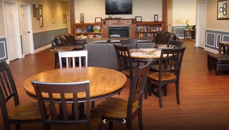 A cozy common area in a senior living facility featuring wooden round tables with chairs, a seating area with brown leather sofas, a fireplace with a wooden mantel, bookshelves filled with books and games, and a wall-mounted flat screen TV. The room has wood flooring and light-colored walls with wainscoting.