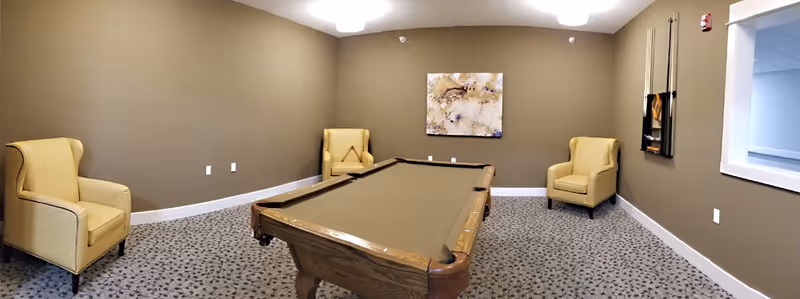 A game room with a pool table in the center, surrounded by three beige armchairs placed against brown walls. A modern abstract painting hangs on the far wall, and a rack with pool cues is mounted on the right wall. The floor is carpeted with a patterned design.