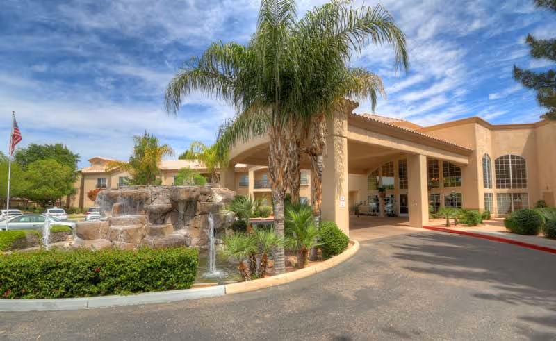 Exterior view of The Montecito Senior Living facility showing a driveway entrance with a covered drop-off area, palm trees, a rock water fountain, and an American flag under a partly cloudy sky.