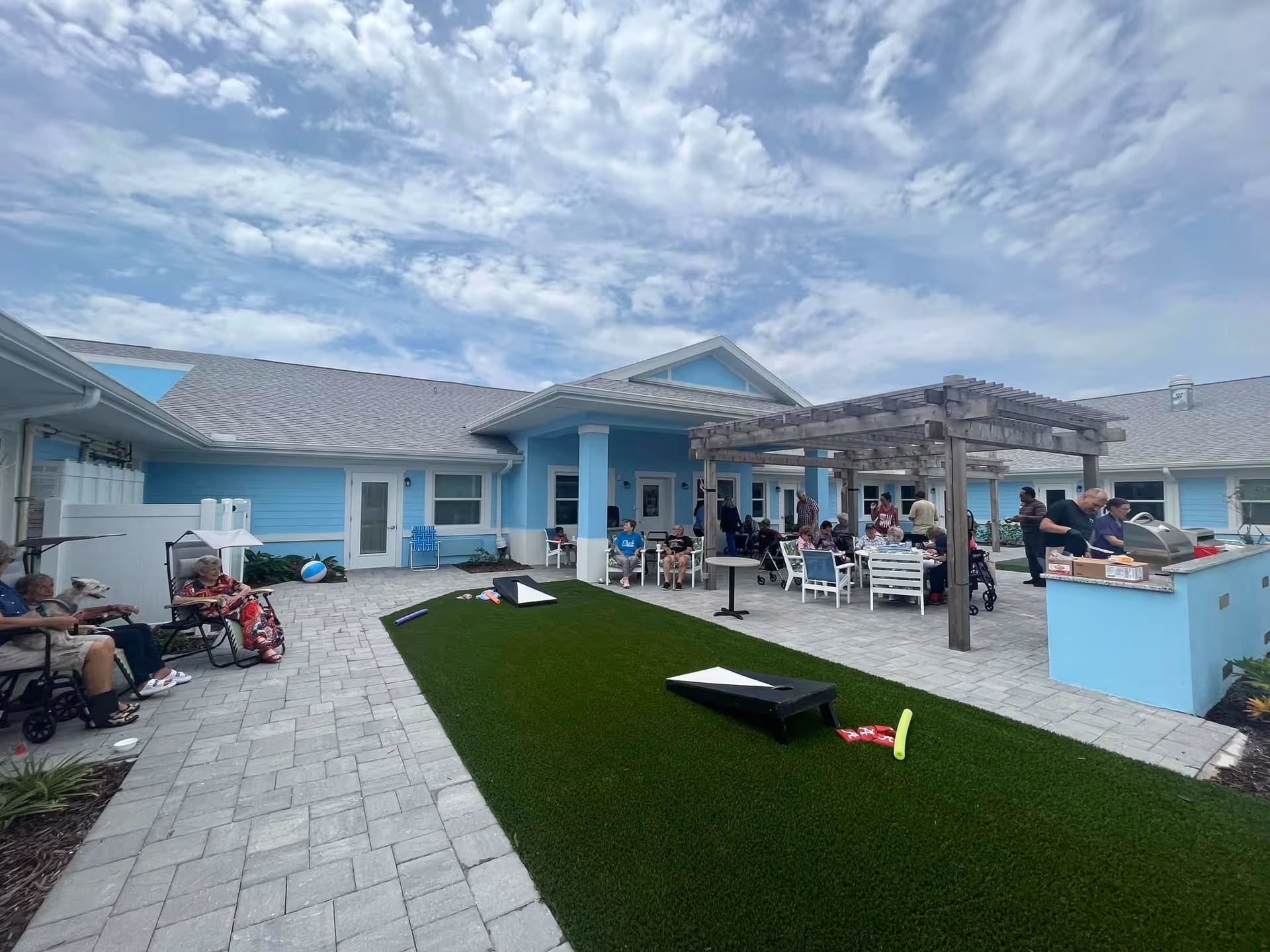 Outdoor courtyard at a blue assisted-living building with seniors and staff gathered under a pergola, seating, a grill area, and lawn games.