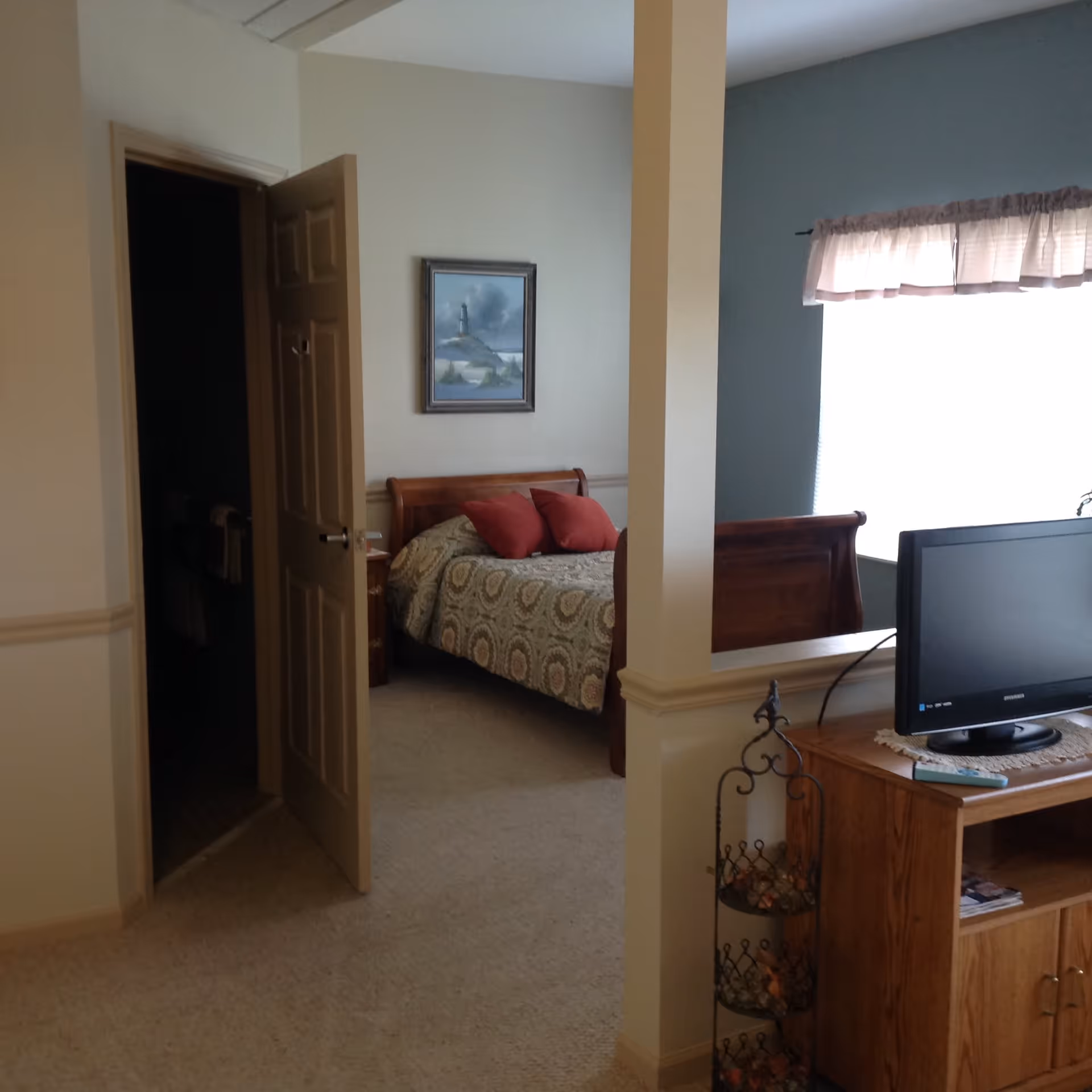 Interior view of a bedroom in Greenbriar Healthcare Center featuring a wooden bed with patterned bedding and red pillows, a framed painting of a lighthouse on the wall, an open door leading to a dark bathroom, a window with sheer curtains, and a wooden TV stand with a flat-screen TV and decorative items.