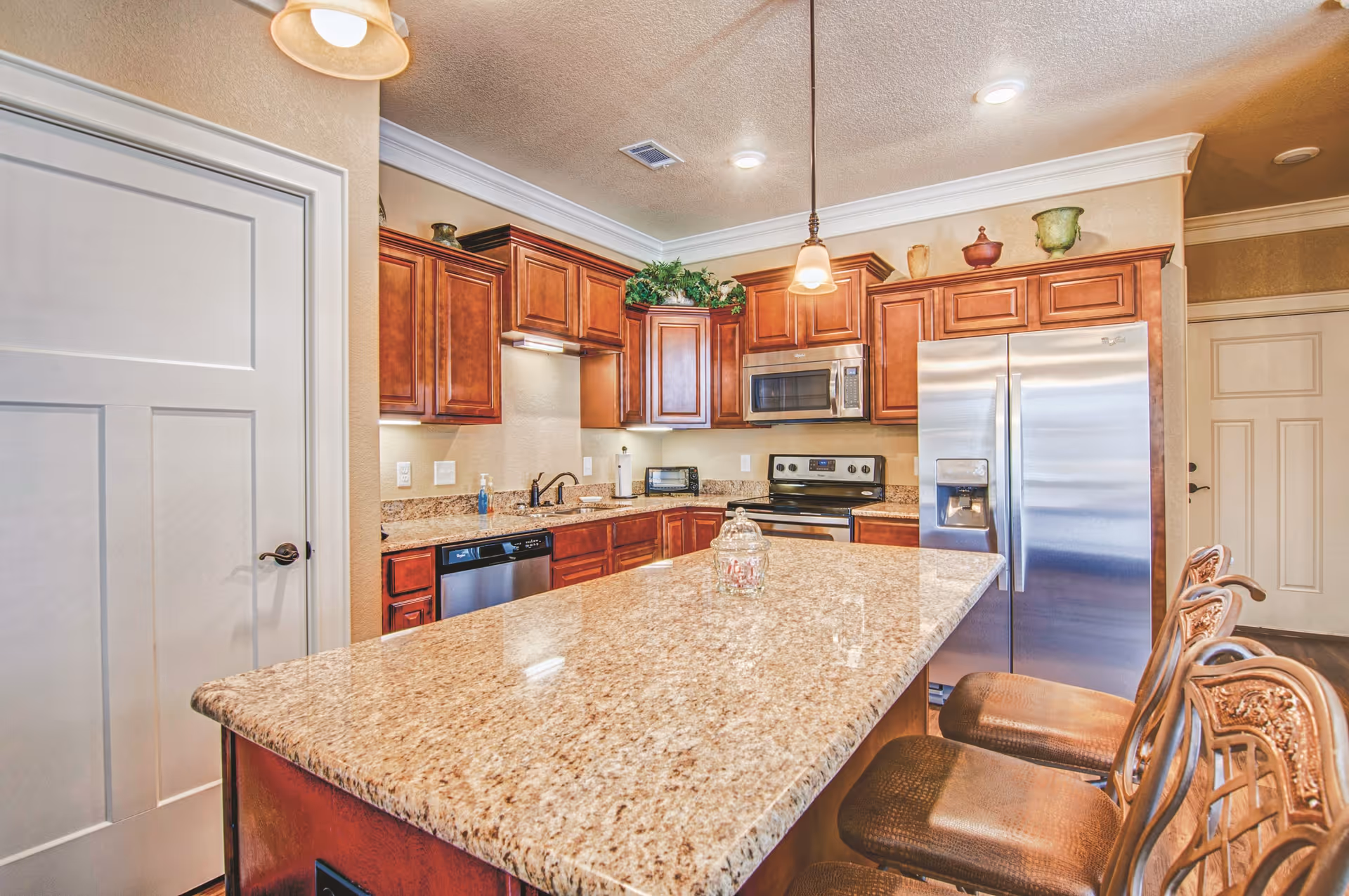 A modern kitchen with granite countertops, wooden cabinets, stainless steel appliances including a refrigerator, microwave, and stove. There is a large kitchen island with three ornate bar stools. The kitchen is well-lit with ceiling lights and pendant lights above the island.