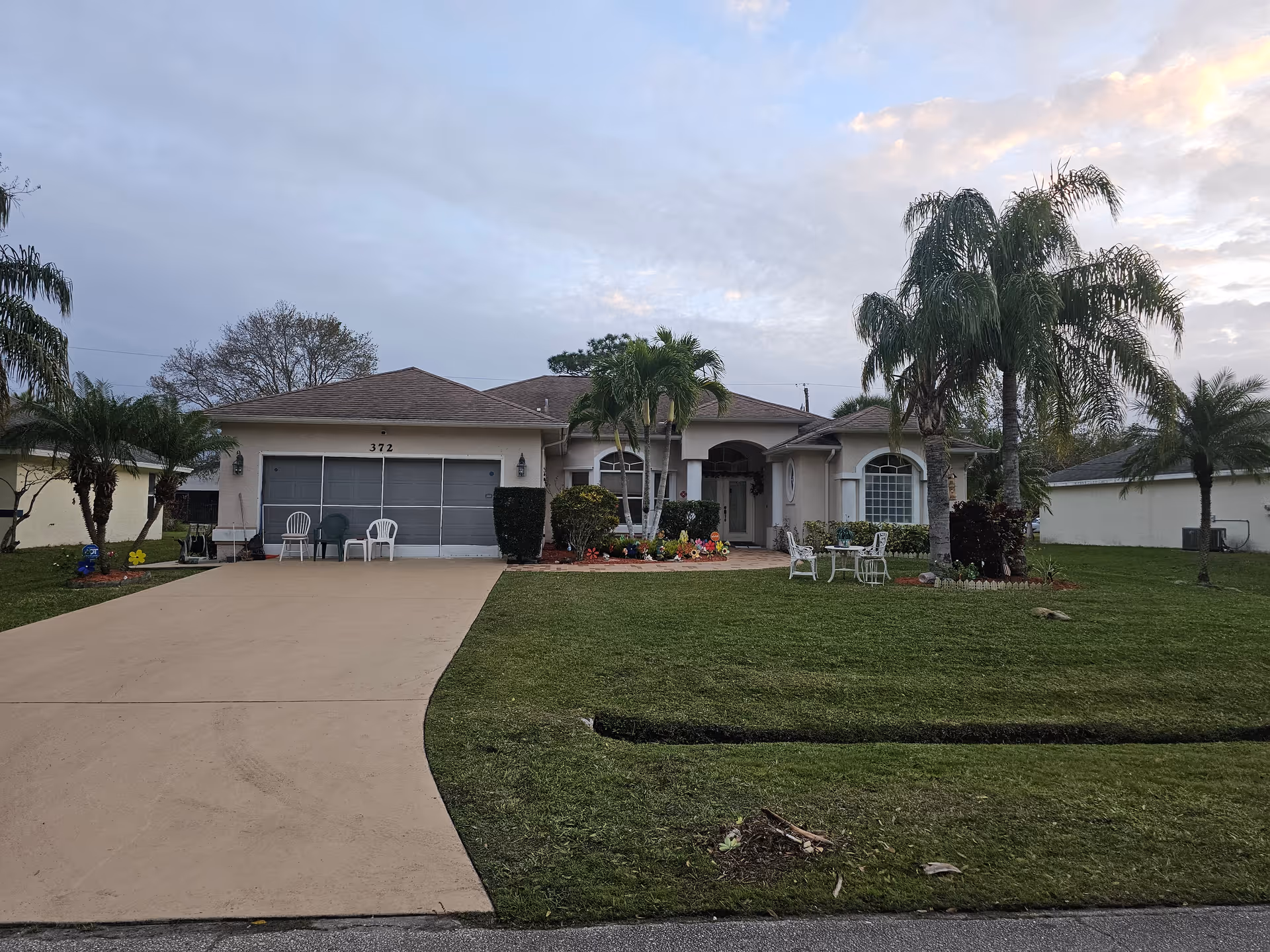 A single-story residential house with a beige exterior and a brown roof. The house has a two-car garage with three chairs placed in front of it. There is a well-maintained lawn with palm trees and a small garden area with flowers near the entrance. A small outdoor seating area with a table and chairs is also visible on the lawn. The sky is partly cloudy during what appears to be early evening or late afternoon.
