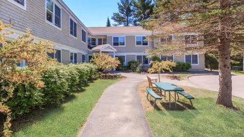 Exterior view of Merry Gardens senior living with a path, trees, and picnic tables.