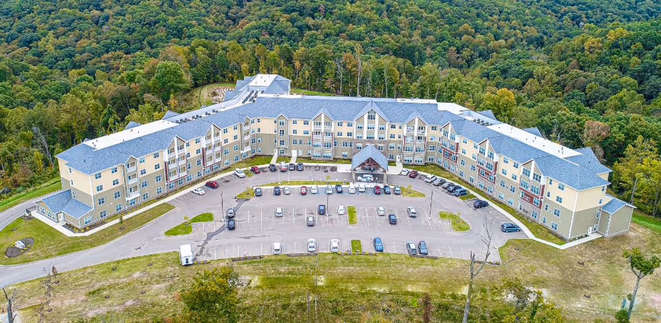 Aerial view of a large senior living facility named Harmony at Southridge, surrounded by dense green forest. The building is U-shaped with multiple floors and a central entrance with a covered drop-off area. There is a parking lot in front with several cars parked and open spaces around the building.