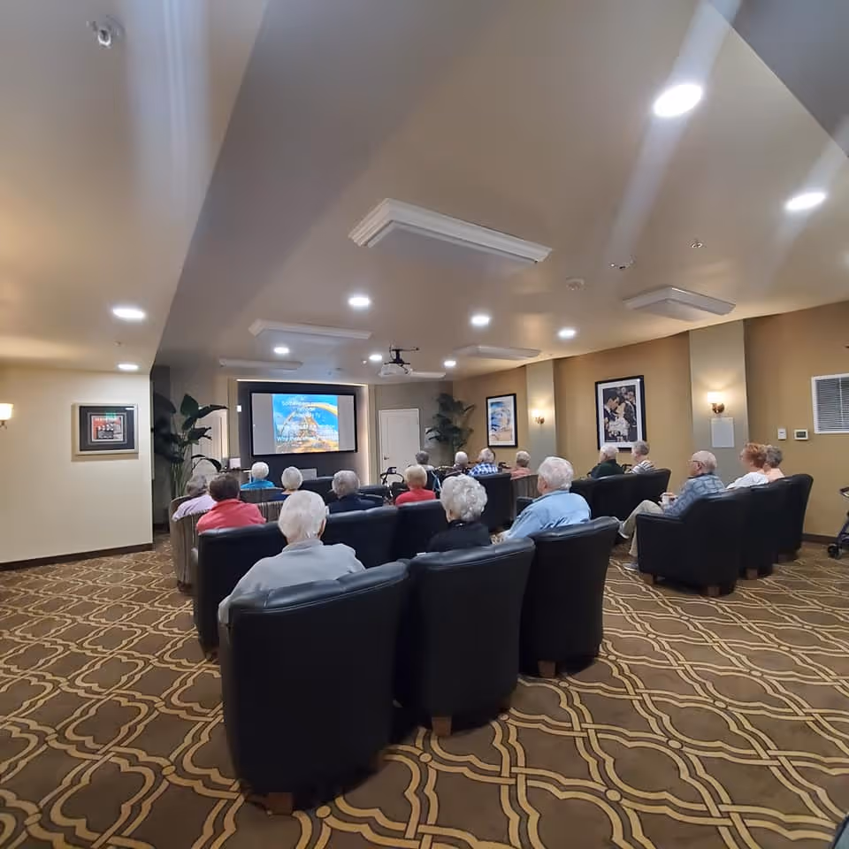 A group of elderly people seated in black armchairs in a carpeted room watching a presentation on a large screen at the front. The room has beige walls with framed pictures and plants, and ceiling lights providing bright illumination.