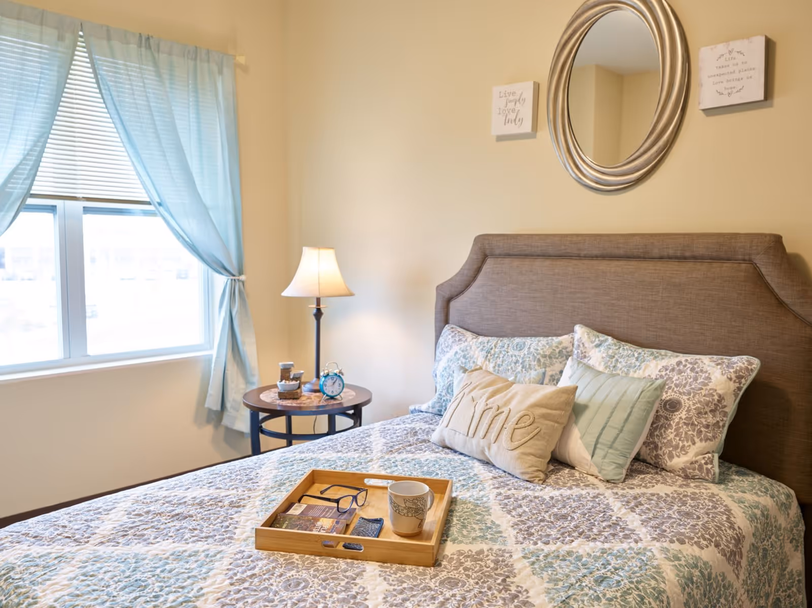 A cozy bedroom with a neatly made bed featuring patterned bedding and multiple pillows, including one with the word 'Mine'. A wooden tray on the bed holds a pair of glasses, a cup, and some reading materials. Next to the bed is a round side table with a lamp, an alarm clock, and some small items. A window with light blue curtains allows natural light into the room. Above the bed is a round mirror and two small framed quotes on the wall.