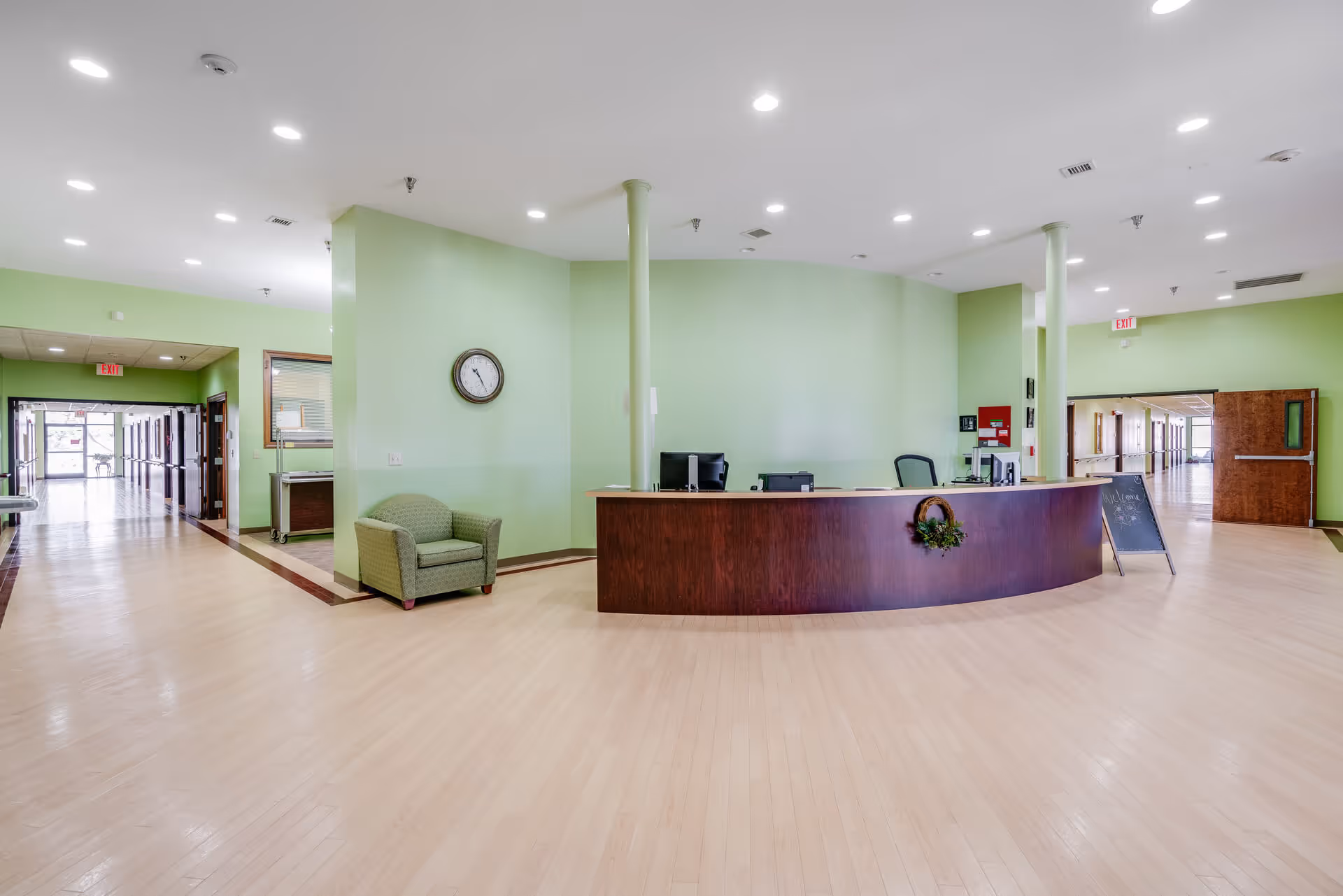 A spacious senior living facility reception area with light green walls and light wood flooring. A curved wooden reception desk is centered with two computer monitors and office chairs behind it. To the left of the desk is a green upholstered armchair and a wall clock. Two long hallways extend from the reception area, with exit signs visible above doors at the end of each hallway. The area is well-lit with recessed ceiling lights.