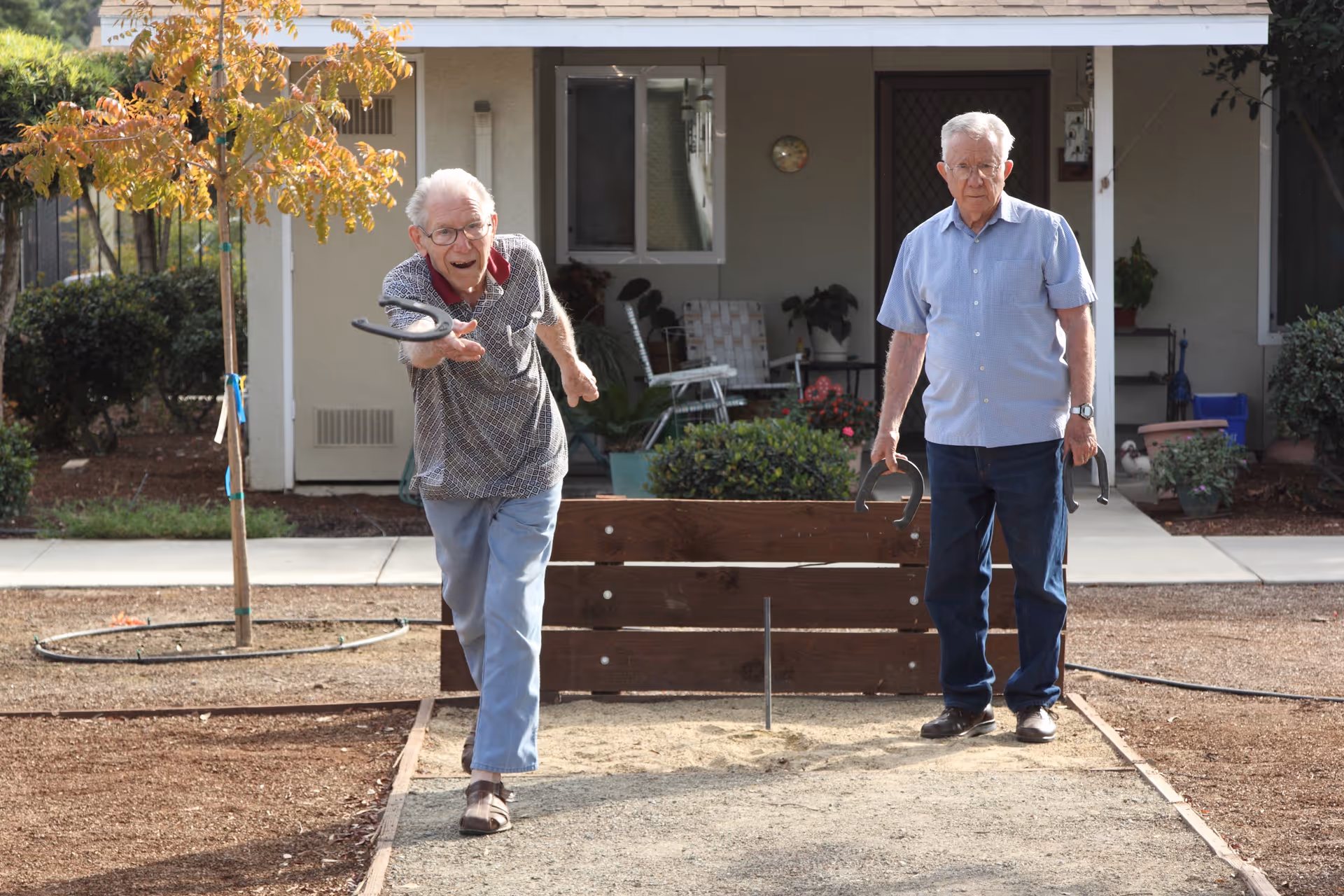 Two elderly men playing horseshoes outdoors in a garden area of a retirement community. One man is in the act of throwing a horseshoe while the other watches, holding two horseshoes. There is a small tree and some bushes in the background, along with a building entrance.