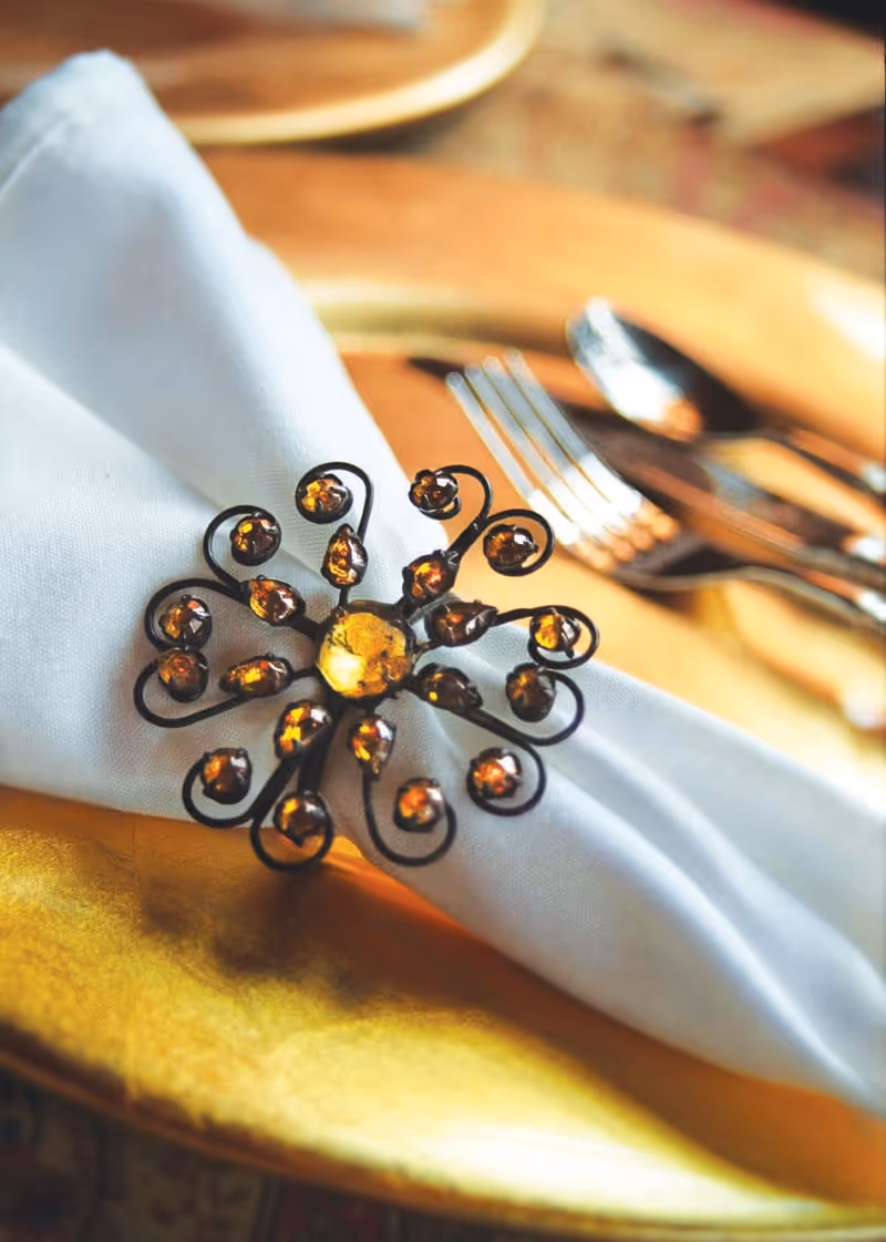 Close-up of a white cloth napkin held by a decorative napkin ring with amber-colored stones, placed on a gold charger plate with silverware in the background.