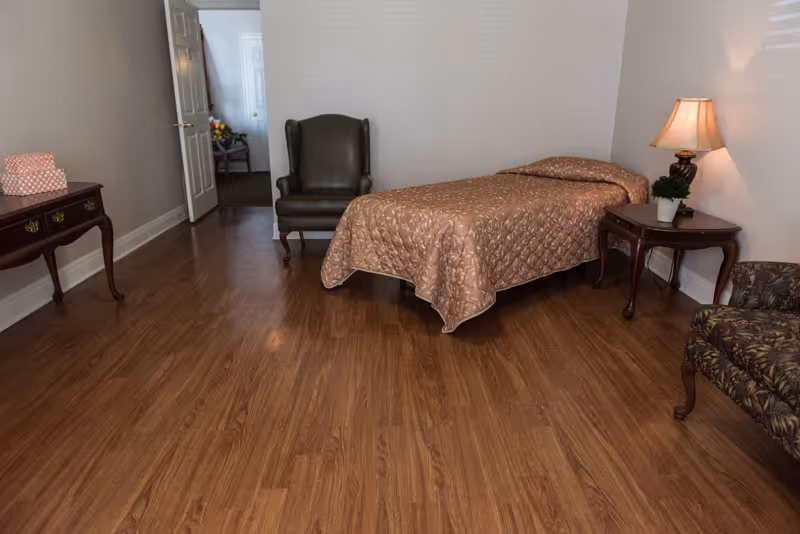 A simple bedroom with a single bed covered in a brown quilted bedspread, a wooden side table with a lamp and a small plant, a green armchair, a floral upholstered chair, and a wooden console table with a pink box on it. The room has wood flooring and white walls, with an open door leading to another room.