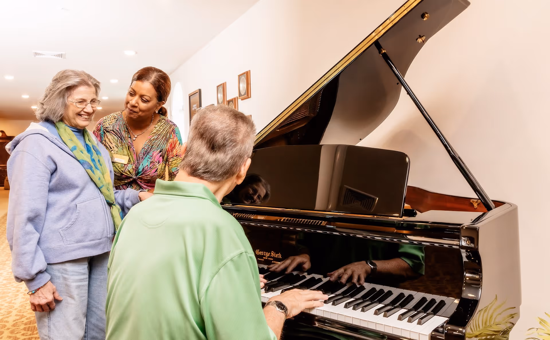 An elderly woman smiling and standing next to a woman in a colorful blouse who is looking at her warmly, while a man in a green shirt plays a grand piano in a well-lit room with framed pictures on the wall.