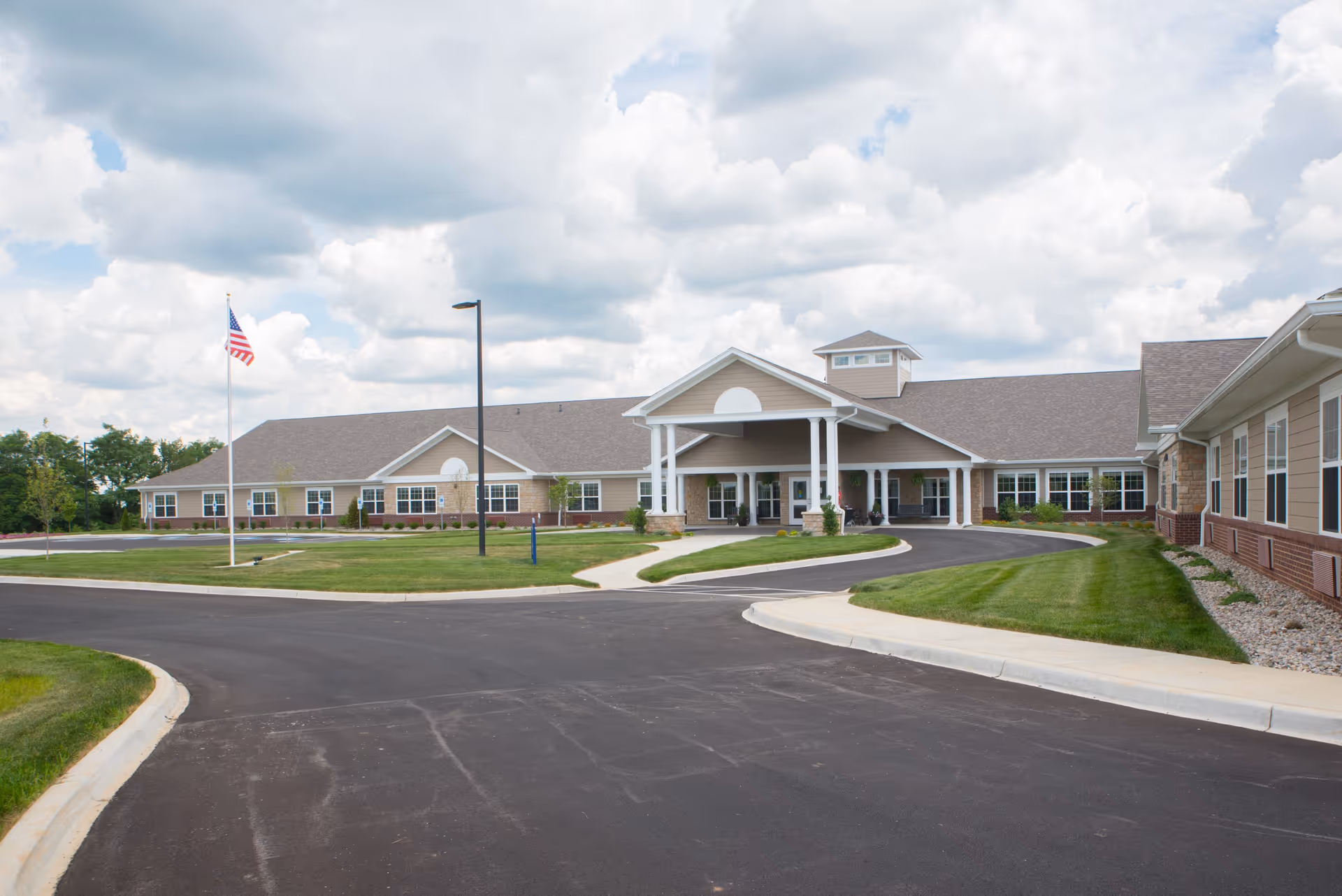 Wide front view of a single-story senior living facility with a covered porte-cochère entrance, flagpole, curved driveway, and landscaped lawn under a cloudy sky.