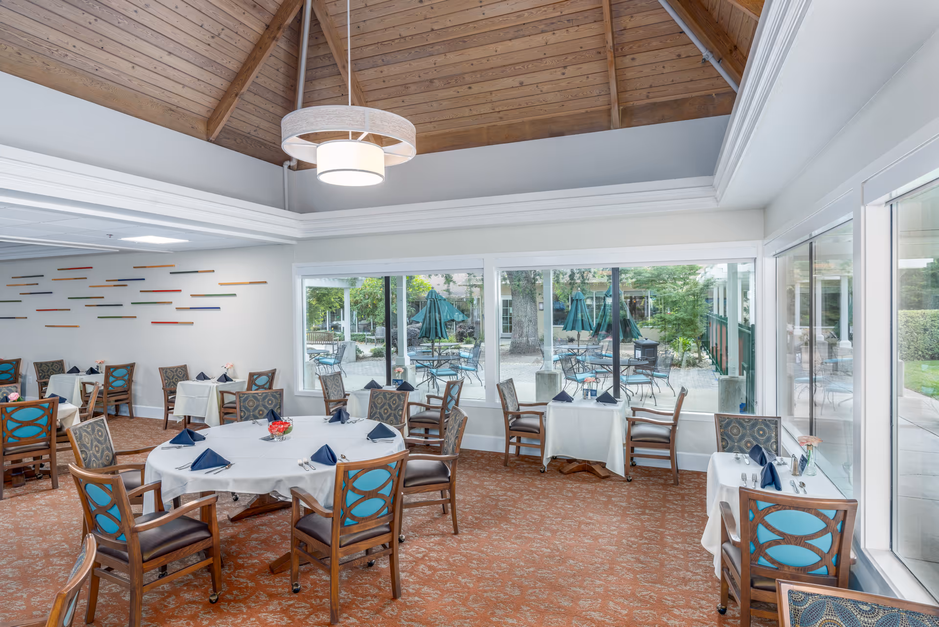 A dining room with round and square tables covered with white tablecloths, each set with navy blue napkins and silverware. The room features wooden chairs with blue patterned cushions, large windows showing an outdoor patio with tables and umbrellas, and a high wooden ceiling with a modern circular light fixture.