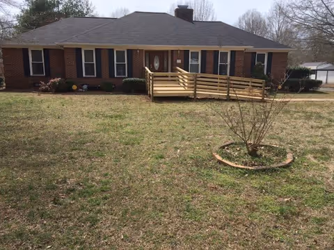 Single-story brick house with a dark shingled roof and multiple windows with black shutters. A wooden wheelchair ramp leads to the front door. The house is surrounded by a large grassy yard with a small circular flower bed containing a leafless shrub. Trees are visible in the background under a cloudy sky.