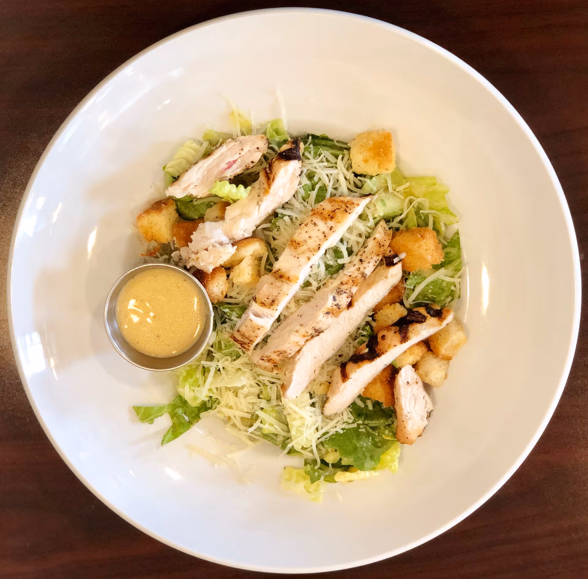 A white bowl containing a Caesar salad with grilled chicken strips, shredded cheese, croutons, and a small metal cup of dressing on the side, placed on a wooden table.