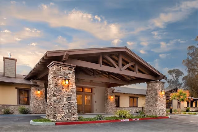 Exterior view of a single-story building with a large covered entrance supported by stone pillars. The building has warm lighting and is surrounded by a paved driveway and some landscaping. The sky is partly cloudy during sunset or sunrise.
