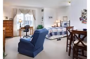 Bright furnished studio bedroom with a bed, blue recliner, desk, dresser and small dining table.