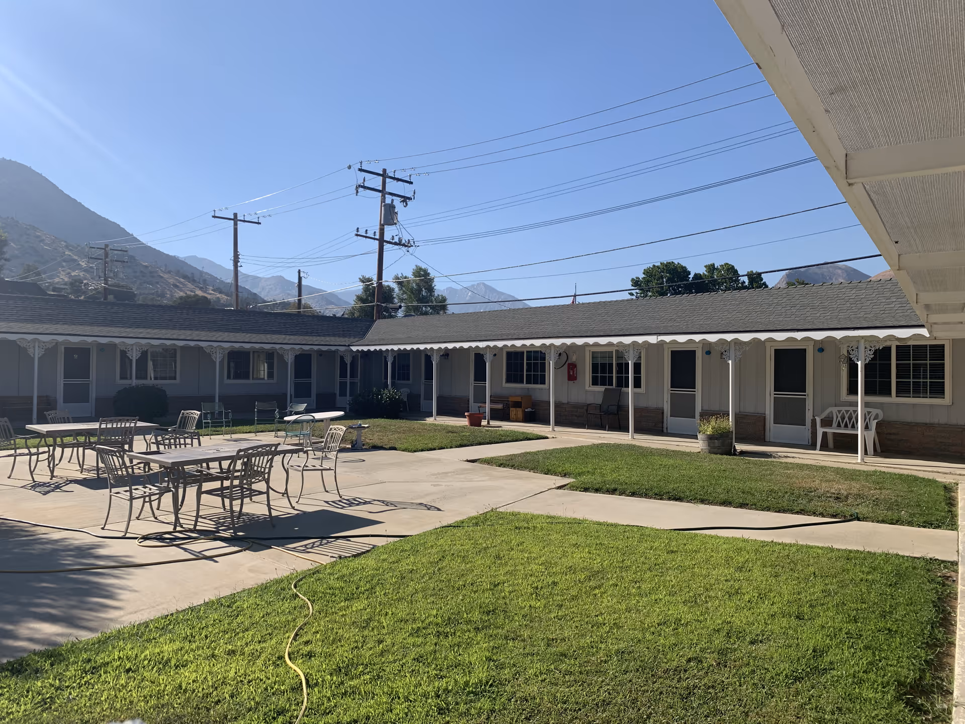 Sunny courtyard of a single-story assisted living building with patio tables and chairs, a grassy lawn, walkways, and mountains in the background.