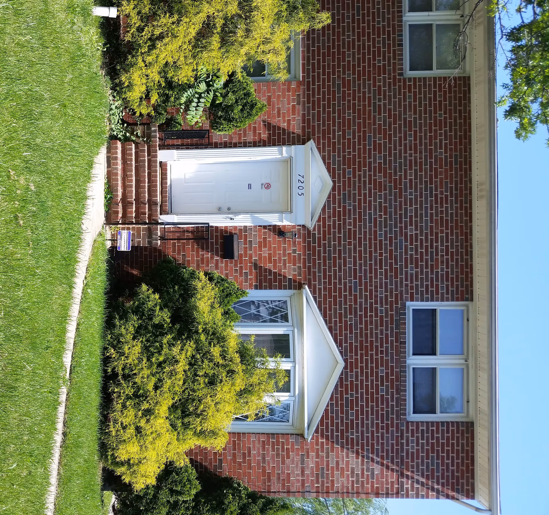 Front exterior view of a red brick building with a white door numbered 7205, two windows, and green bushes and grass in front.