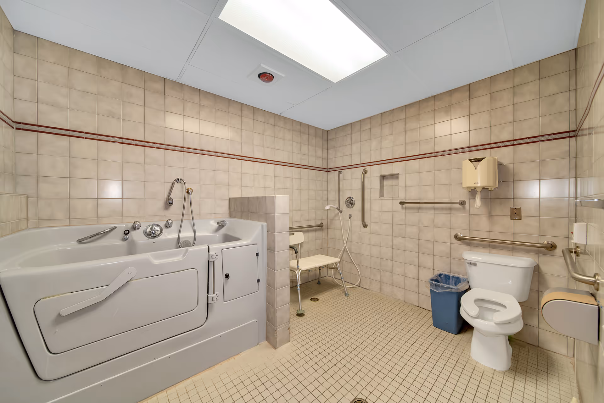 A spacious accessible bathroom with beige tiled walls and floor. The room features a walk-in bathtub with a door and handrails, a shower area with a foldable chair and handheld showerhead, a toilet with grab bars on both sides, a wall-mounted soap dispenser, and a blue trash bin. The ceiling has a large light panel.