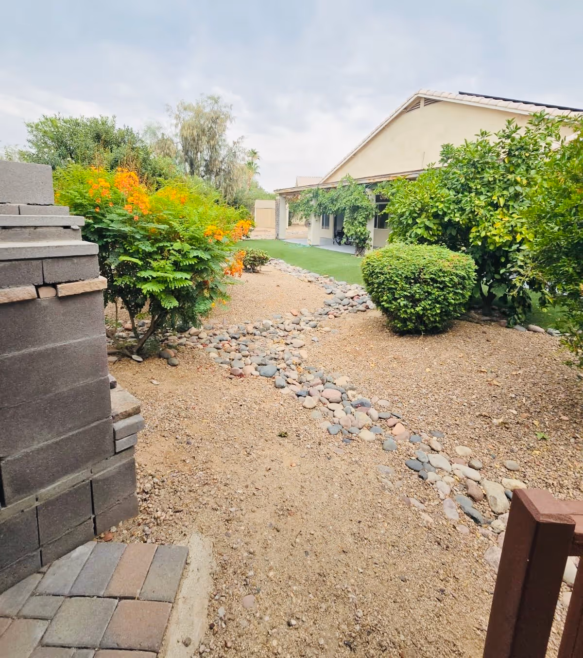 Gravel backyard with a rock-lined dry creek, shrubs and flowering plants leading to a covered patio of a single-story house.