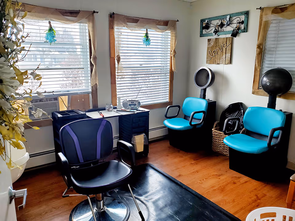 A small salon room with two turquoise salon chairs and one black salon chair with purple accents. The room has wooden floors and three windows with white blinds and beige curtains. There is a small table with a radio and some papers between the windows. On the wall, there is a decorative piece with the word 'love' and a coat rack with some items hanging. A basket with folded chairs is in the corner.
