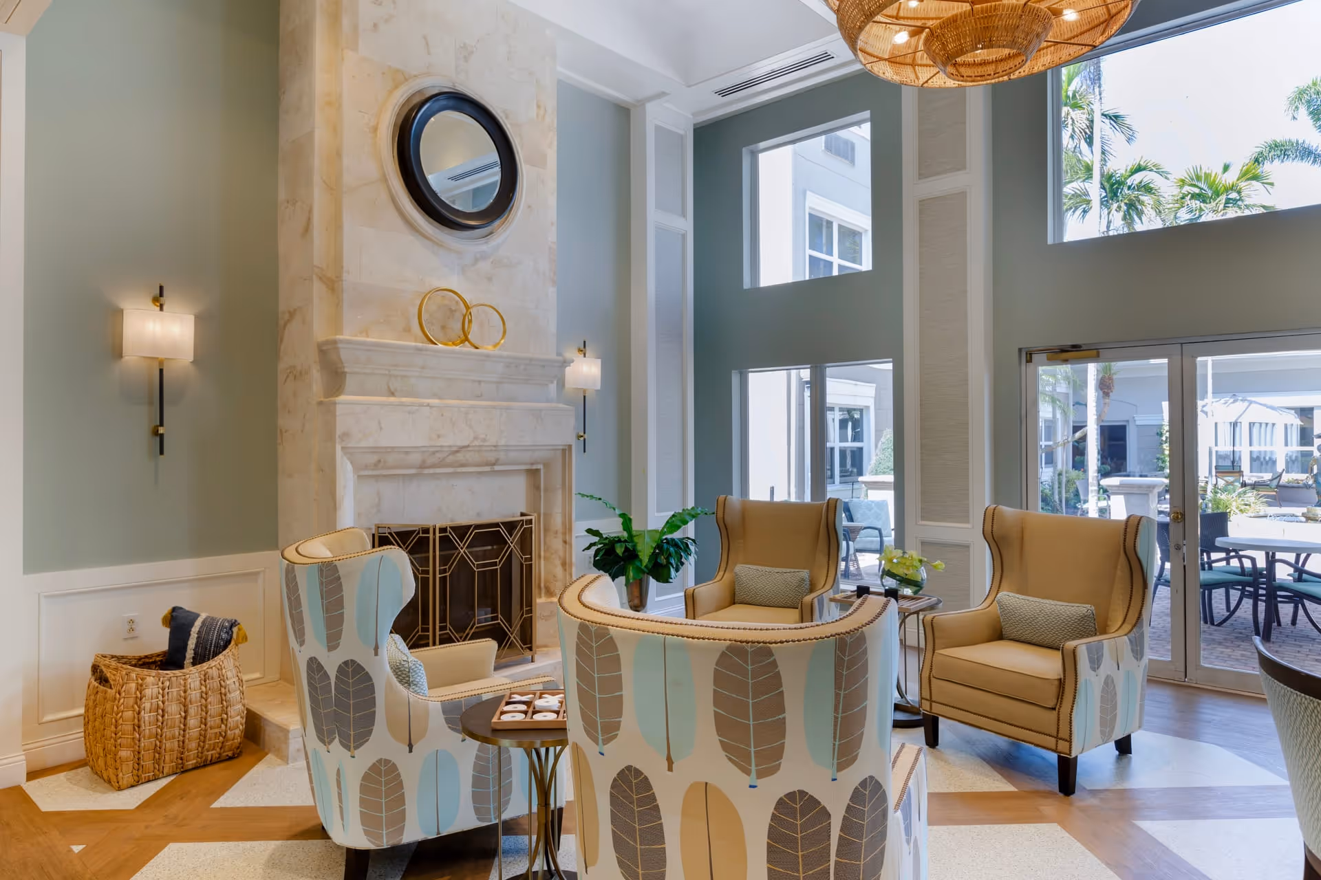 A bright and airy living room area with a tall marble fireplace, two beige armchairs, and two patterned armchairs with leaf designs. There is a round mirror and decorative rings above the fireplace, wall sconces on either side, a woven basket with pillows, and large windows letting in natural light with a view of an outdoor patio with tables and chairs.