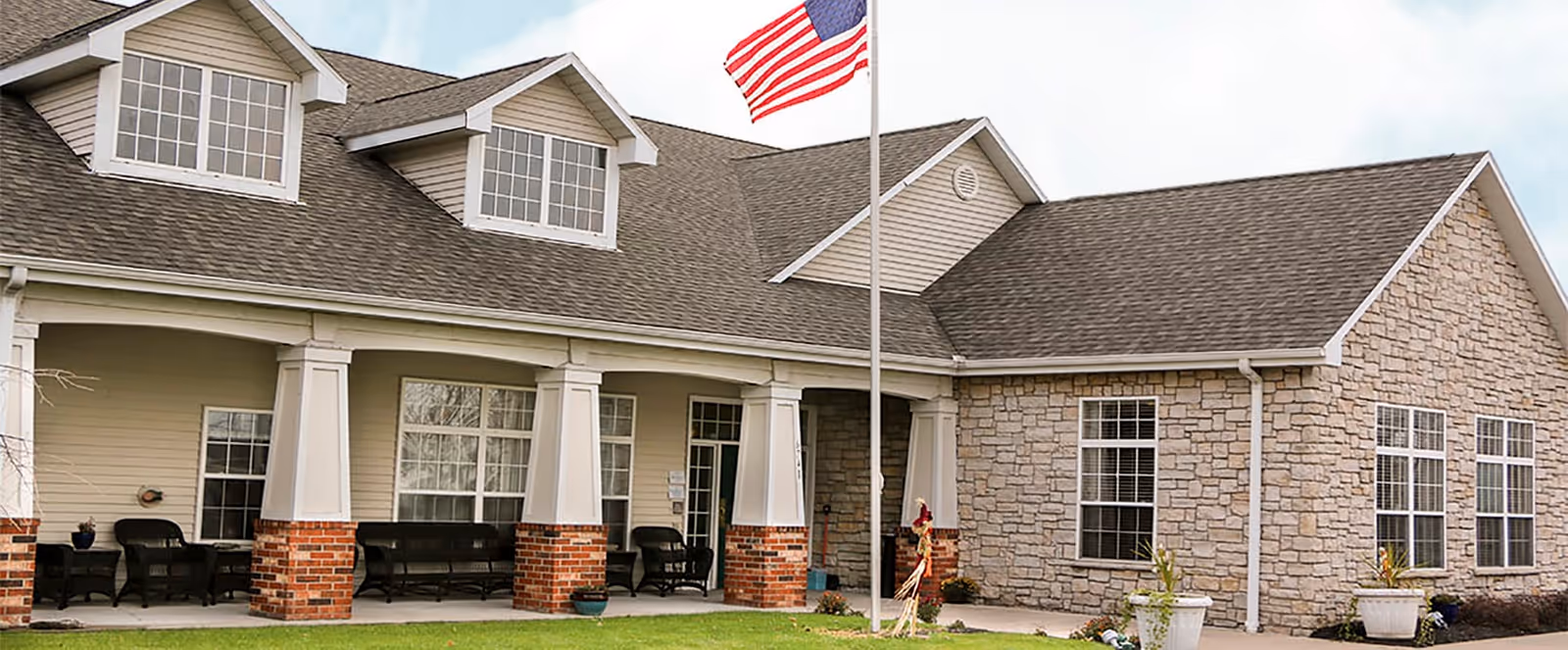 Exterior view of a senior living facility building with a covered porch supported by white columns with brick bases. The building has beige siding and stone walls, multiple windows, and dormer windows on the roof. An American flag is flying on a flagpole in front of the building.