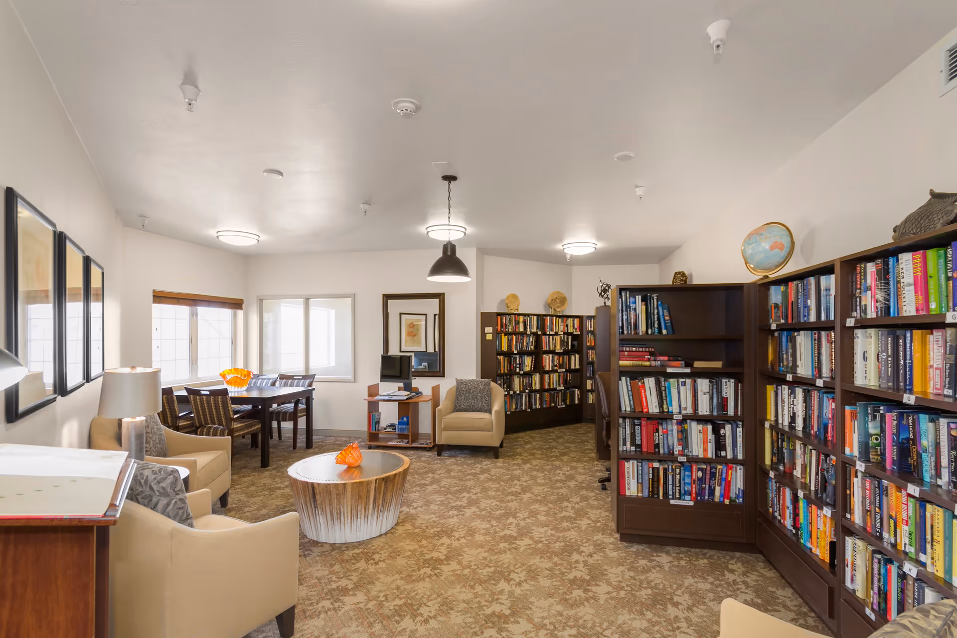 A cozy library and sitting area in a senior living facility with bookshelves filled with books along the right wall, comfortable beige armchairs, a round wooden coffee table with an orange decorative piece, and a dining table with chairs near windows on the left side. The room is well-lit with ceiling lights and has carpeted flooring.
