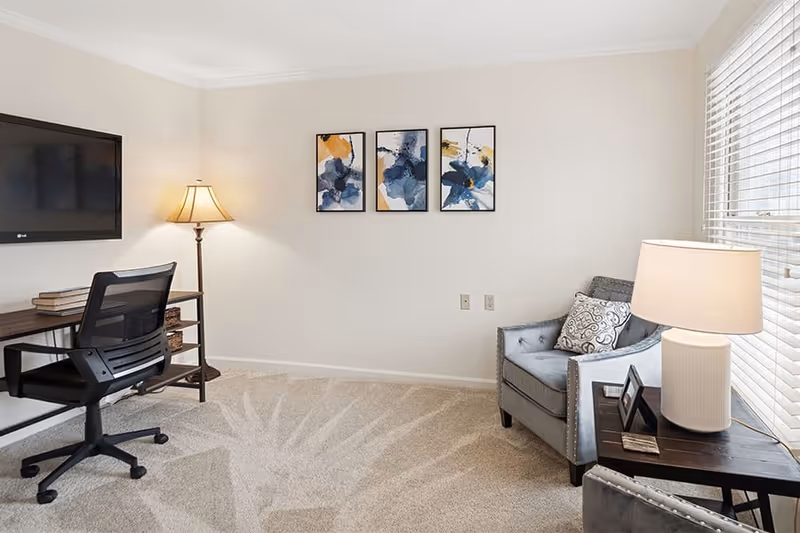 A cozy interior room with a gray armchair and a side table with a lamp and picture frame near a window with blinds. On the opposite wall, there is a desk with a black office chair, a floor lamp, and a wall-mounted TV. Three abstract art pieces are hung on the wall above the carpeted floor.